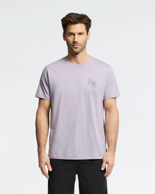 A man with short brown hair wears the Psycho Bunny MENS ELLIOT FRONT BACK GRAPHIC TEE in light purple, made of Pima cotton, and black shorts against a plain white background. The regular fit tee features a small graphic print on the upper left side.