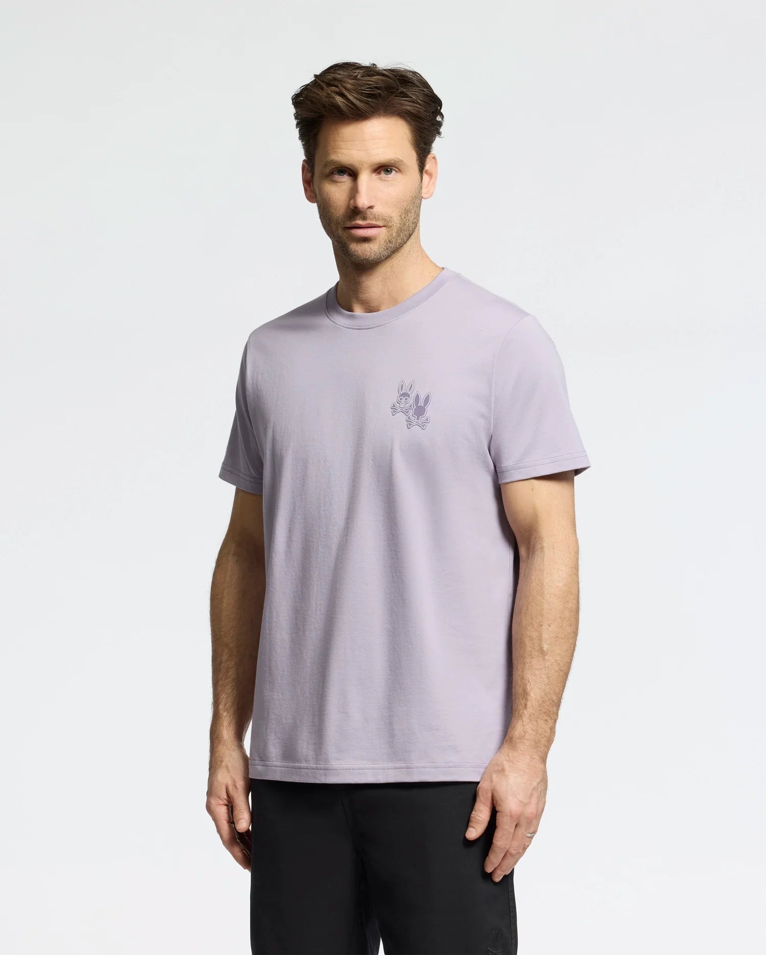 A man in a Psycho Bunny light purple "MENS ELLIOT FRONT BACK GRAPHIC TEE - B6U605F200" stands against a plain white background. The regular fit shirt complements his short brown hair and dark pants.