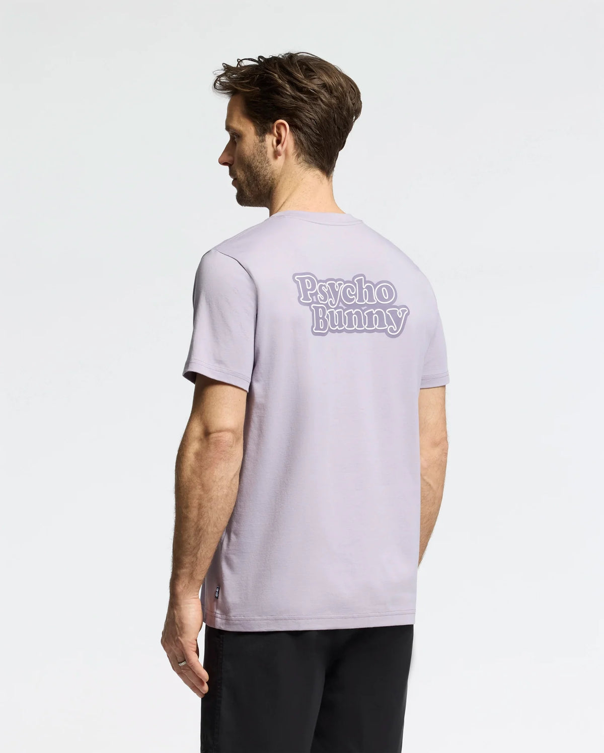 A man with short dark hair stands against a plain white background, wearing Psycho Bunny's MENS ELLIOT FRONT BACK GRAPHIC TEE - B6U605F200 in light gray Pima cotton, featuring "Psycho Bunny" in stylized letters on the back. He pairs it with black pants.