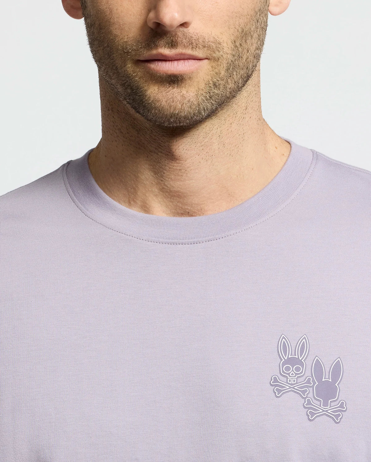 A man sports the Psycho Bunny MENS ELLIOT FRONT BACK GRAPHIC TEE in light purple, featuring a small chest logo with two bunny heads and crossed bones. Its regular fit boosts comfort, focusing attention on the unique design against its plain background.
