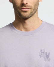 A man sports the Psycho Bunny MENS ELLIOT FRONT BACK GRAPHIC TEE in light purple, featuring a small chest logo with two bunny heads and crossed bones. Its regular fit boosts comfort, focusing attention on the unique design against its plain background.