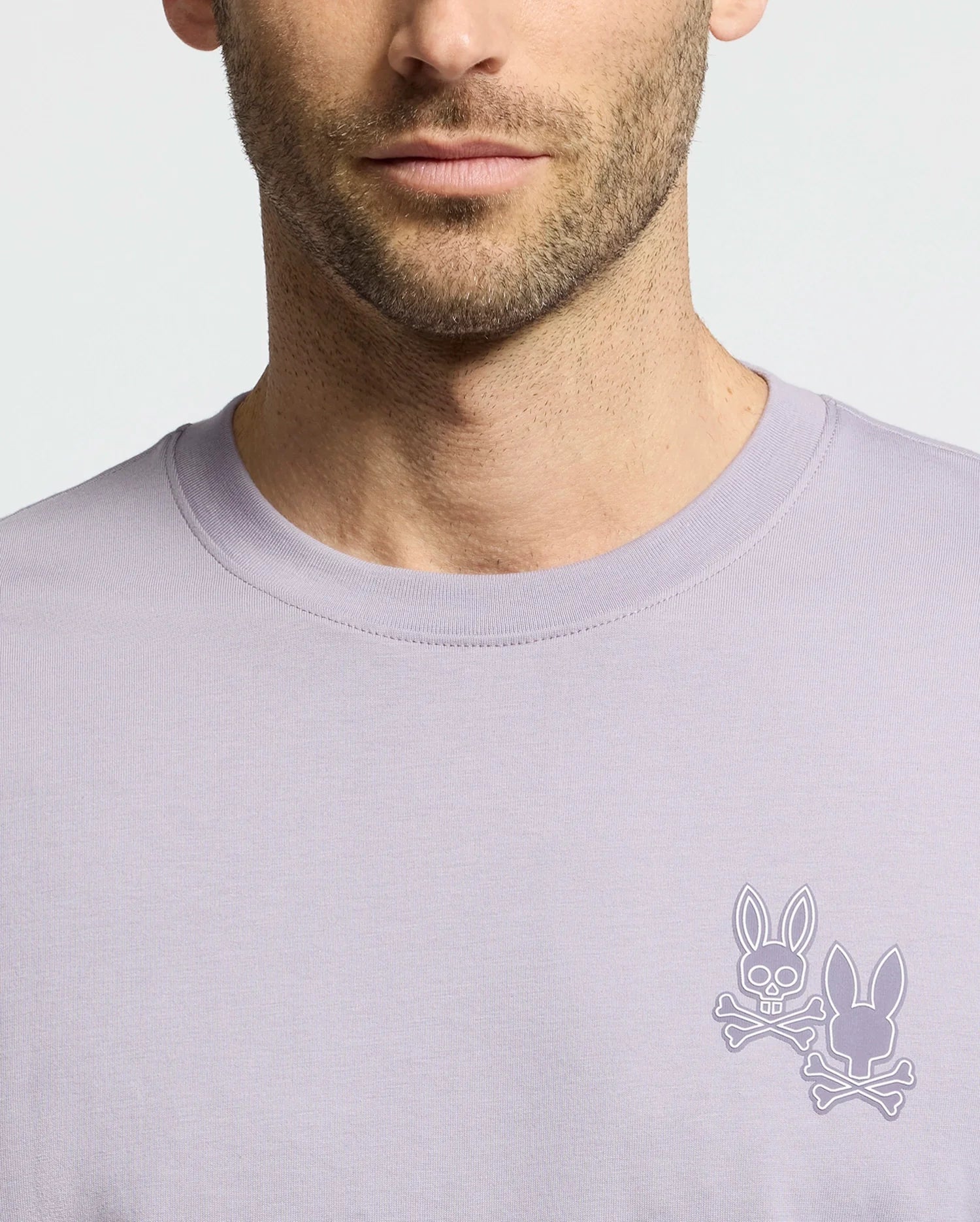 A man sports the Psycho Bunny MENS ELLIOT FRONT BACK GRAPHIC TEE in light purple, featuring a small chest logo with two bunny heads and crossed bones. Its regular fit boosts comfort, focusing attention on the unique design against its plain background.