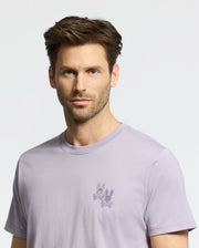 A man with short brown hair and a beard wears a light purple Psycho Bunny MENS ELLIOT FRONT BACK GRAPHIC TEE made from Pima cotton, featuring an embroidered design of two cartoon rabbits on the chest against a plain light-colored background.