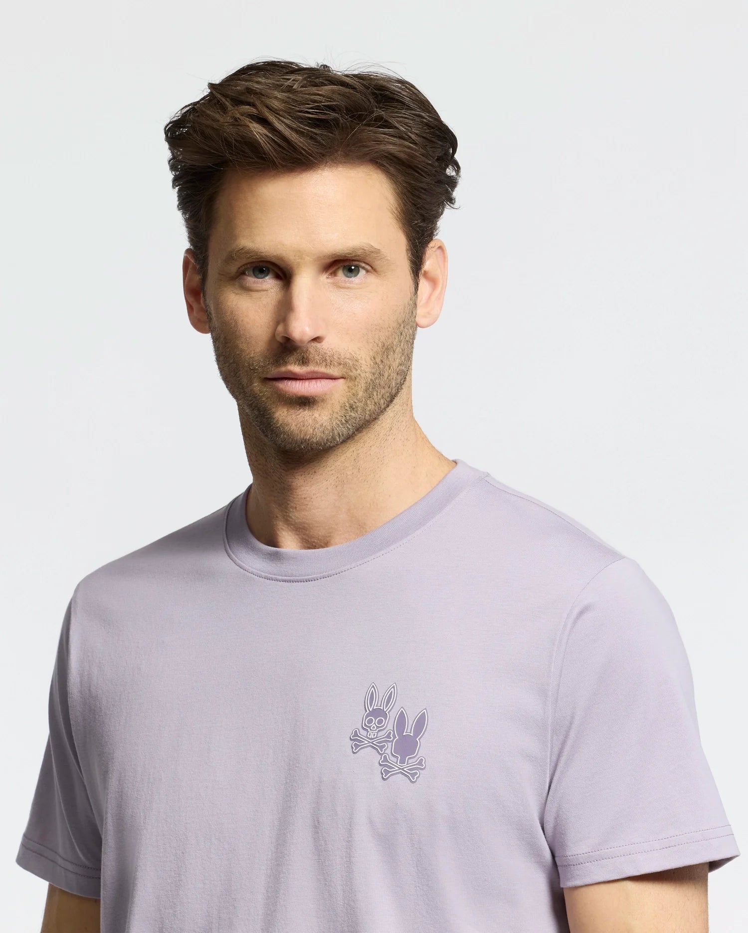 A man with short brown hair and a beard wears a light purple Psycho Bunny MENS ELLIOT FRONT BACK GRAPHIC TEE made from Pima cotton, featuring an embroidered design of two cartoon rabbits on the chest against a plain light-colored background.