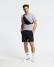 A man confidently stands in a Psycho Bunny MENS ELLIOT FRONT BACK GRAPHIC TEE - B6U605F200, black shorts, white socks, and sneakers. With hands in pockets and a black crossbody bag against a plain white background, he exudes effortless style and comfort.
