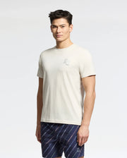 A man stands against a neutral background wearing Psycho Bunny's MENS ELLIOT FRONT BACK GRAPHIC TEE in light beige Pima cotton with a chest graphic, paired with regular fit dark patterned shorts.