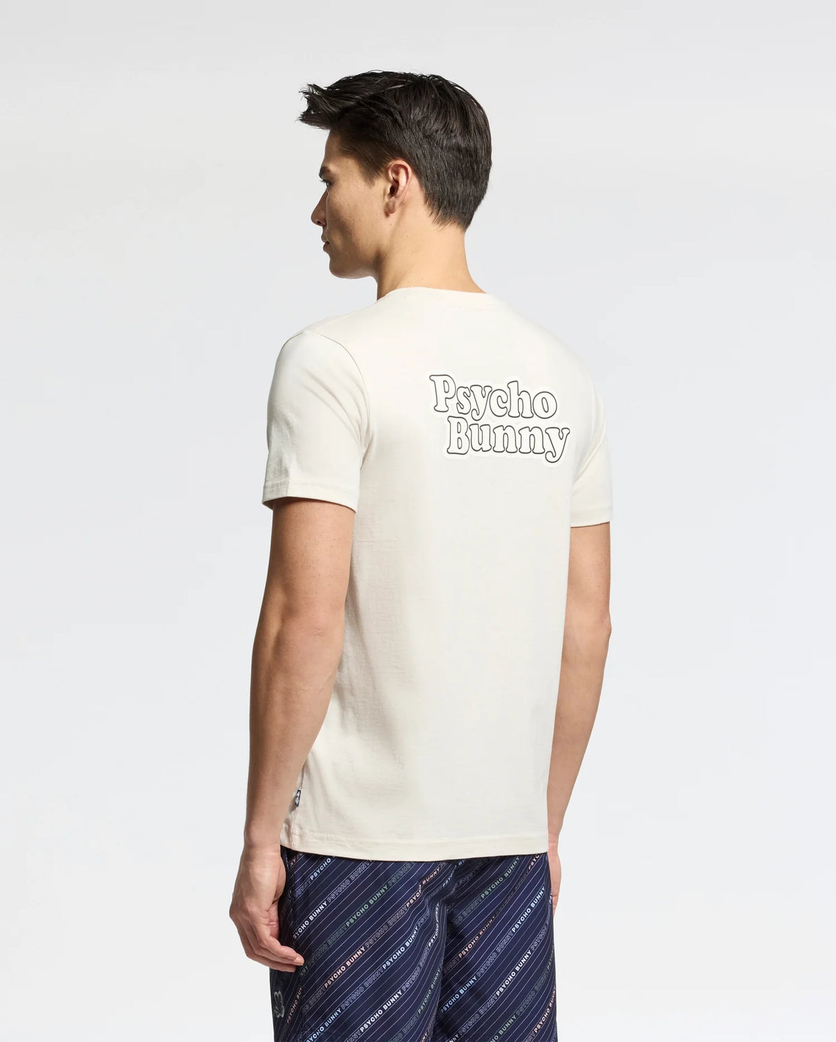 A man with short dark hair models the Psycho Bunny Men's Elliot Front Back Graphic Tee in white Pima cotton, featuring bold "Psycho Bunny" prints on the back. He pairs it with patterned shorts against a light background, offering a regular fit.