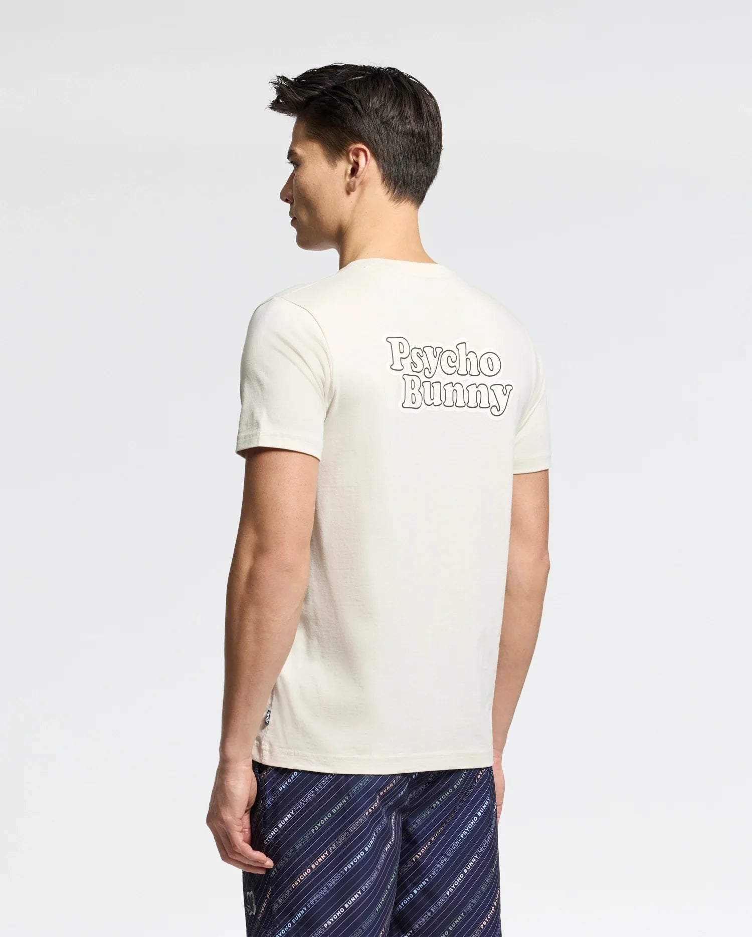 A man with short dark hair models the Psycho Bunny Men's Elliot Front Back Graphic Tee in white Pima cotton, featuring bold "Psycho Bunny" prints on the back. He pairs it with patterned shorts against a light background, offering a regular fit.