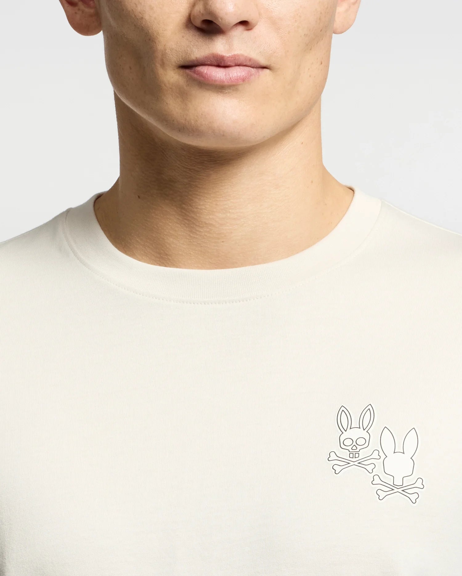 A person wearing a Psycho Bunny MENS ELLIOT FRONT BACK GRAPHIC TEE, a light-colored Pima cotton T-shirt featuring two cartoon-style rabbit head logos with crossed bones on the chest. The regular fit design complements the plain, neutral background perfectly.