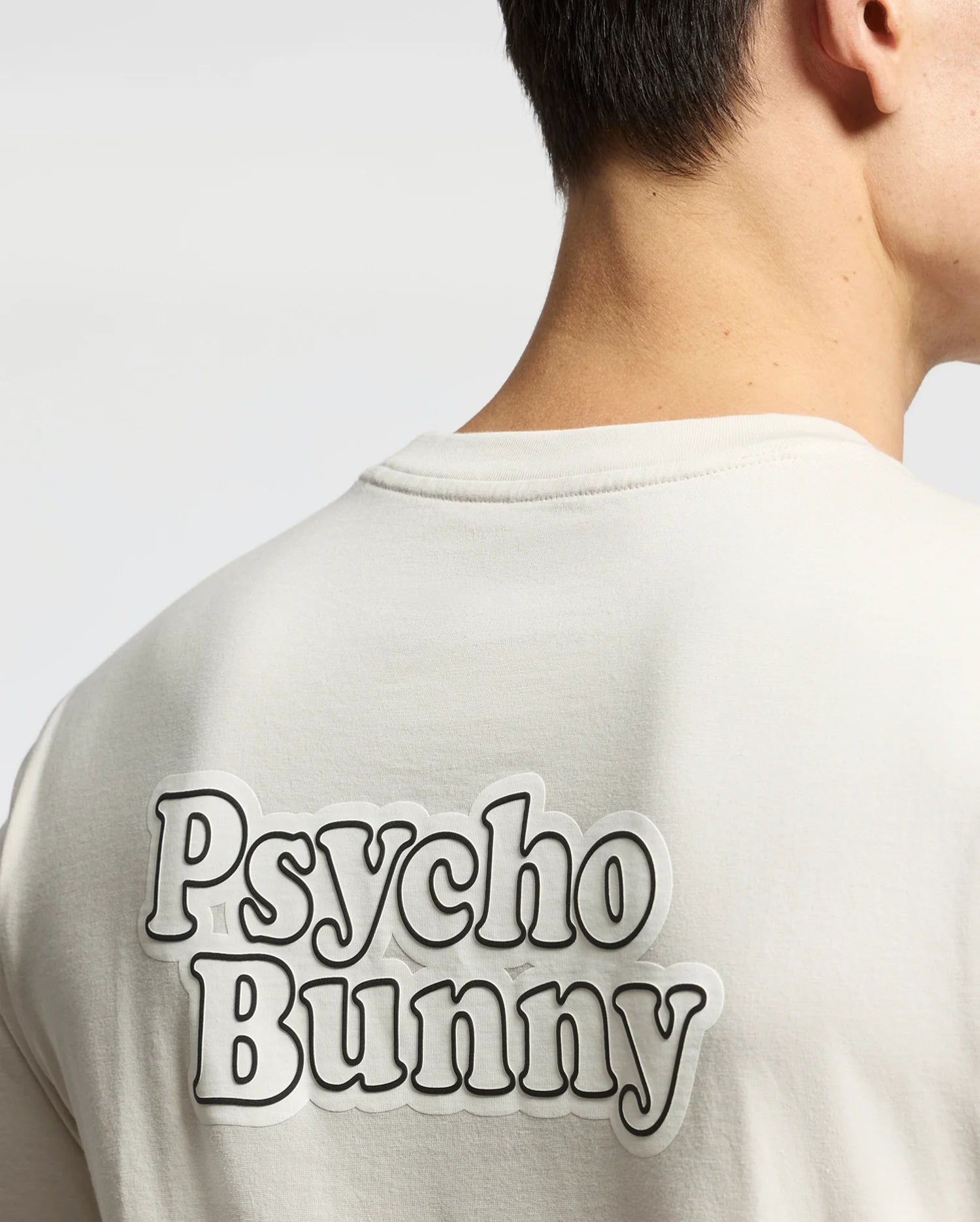 A person with short dark hair dons the MENS ELLIOT FRONT BACK GRAPHIC TEE - B6U605F200 by Psycho Bunny. Its regular fit and bold, outlined "Psycho Bunny" text on the upper back enhance their sleek style.