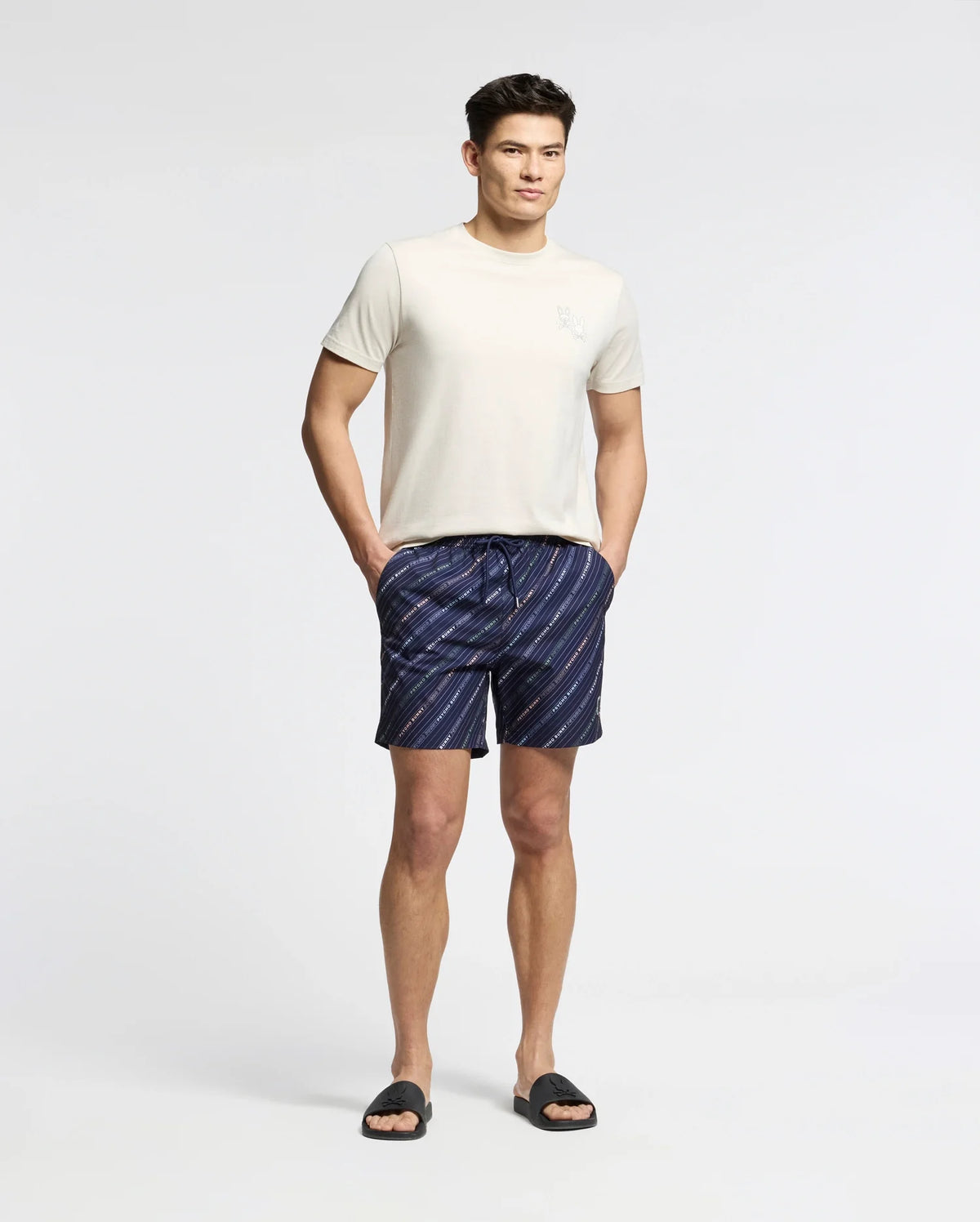 A person stands against a plain background, wearing a Psycho Bunny's MENS ELLIOT FRONT BACK GRAPHIC TEE, dark blue striped shorts with hands in the pockets, and black slide sandals.