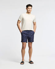 A person stands against a plain background, wearing a Psycho Bunny's MENS ELLIOT FRONT BACK GRAPHIC TEE, dark blue striped shorts with hands in the pockets, and black slide sandals.