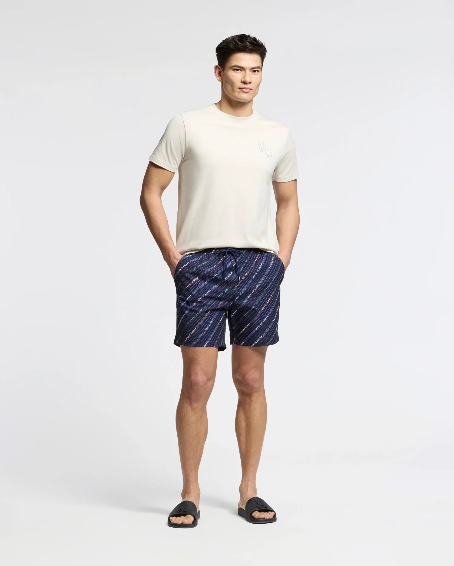 A person stands against a plain background, wearing a Psycho Bunny's MENS ELLIOT FRONT BACK GRAPHIC TEE, dark blue striped shorts with hands in the pockets, and black slide sandals.