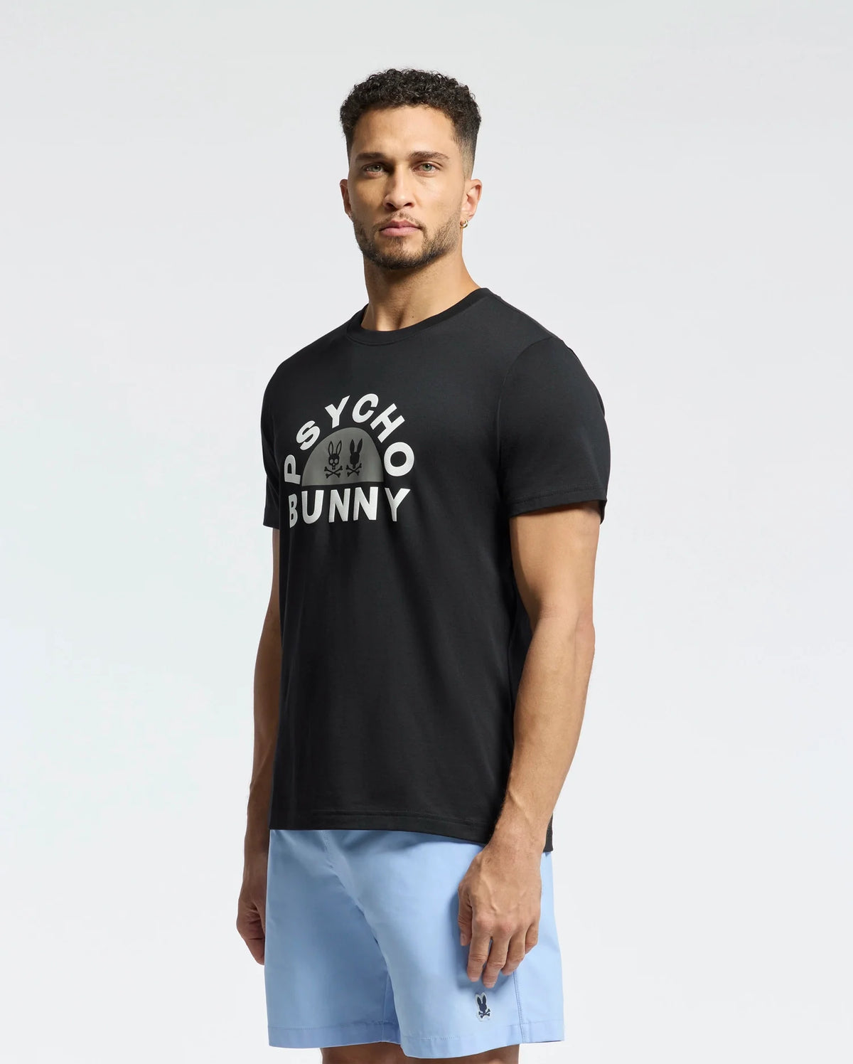 A man stands against a plain background, wearing the Psycho Bunny "MENS GOLDIE GRAPHIC TEE - B6U606F200," made from luxurious Peruvian Pima cotton, and light blue shorts. The black tee showcases a unique Bunny graphic with "Psycho Bunny" above it.