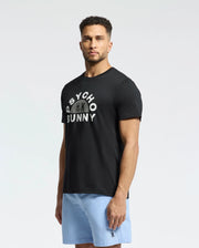 A man stands against a plain background, wearing the Psycho Bunny "MENS GOLDIE GRAPHIC TEE - B6U606F200," made from luxurious Peruvian Pima cotton, and light blue shorts. The black tee showcases a unique Bunny graphic with "Psycho Bunny" above it.