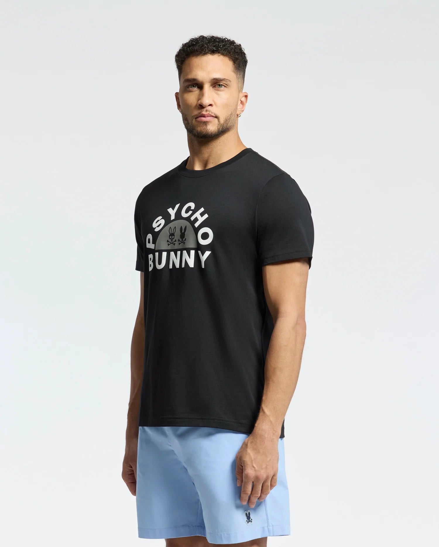 A man stands against a plain background, wearing the Psycho Bunny "MENS GOLDIE GRAPHIC TEE - B6U606F200," made from luxurious Peruvian Pima cotton, and light blue shorts. The black tee showcases a unique Bunny graphic with "Psycho Bunny" above it.