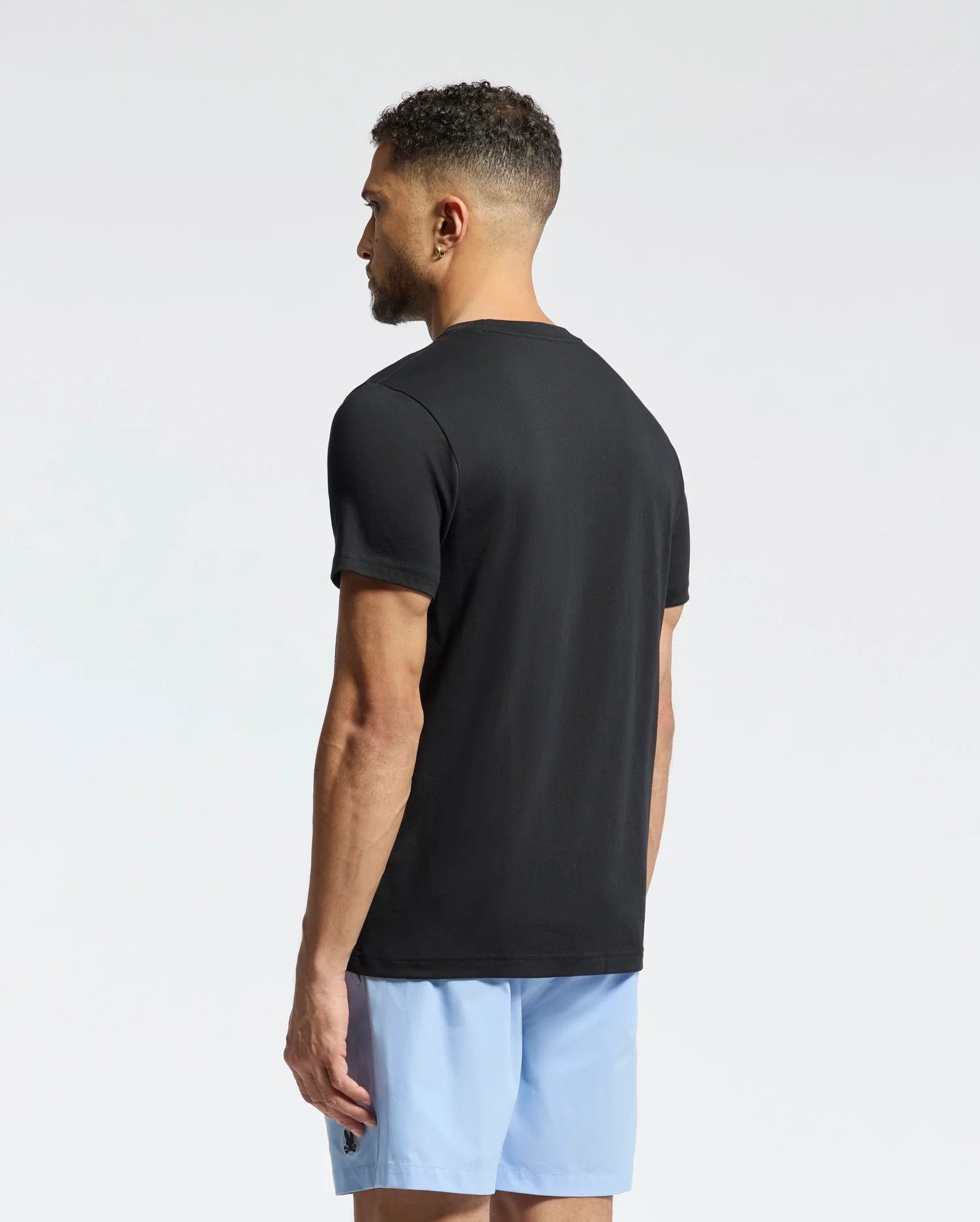 A man with short curly hair, facing away from the camera against a white background, wears a Psycho Bunny MENS GOLDIE GRAPHIC TEE - B6U606F200 in black paired with light blue shorts. Made from Peruvian Pima cotton, the tee offers a casual yet refined appearance.