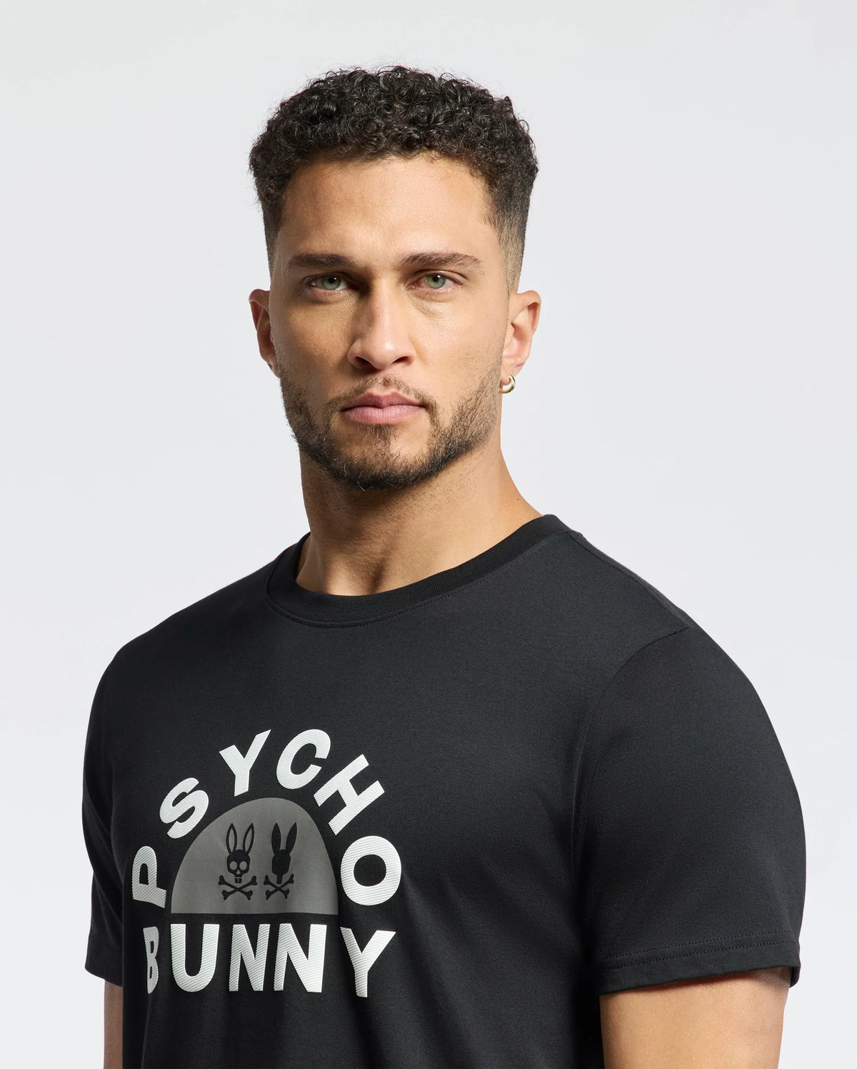 A man with short curly hair, a beard, and hoop earrings wears a Psycho Bunny Mens Goldie Graphic Tee - B6U606F200 against a plain white background. The tee is made of luxurious Peruvian Pima cotton and features a distinctive bunny graphic.