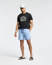 A man stands against a plain backdrop, wearing a black cap and the MENS GOLDIE GRAPHIC TEE by Psycho Bunny, crafted from premium Pima cotton. He's paired it with light blue shorts and black slide sandals, hands by his sides and looking slightly downward.