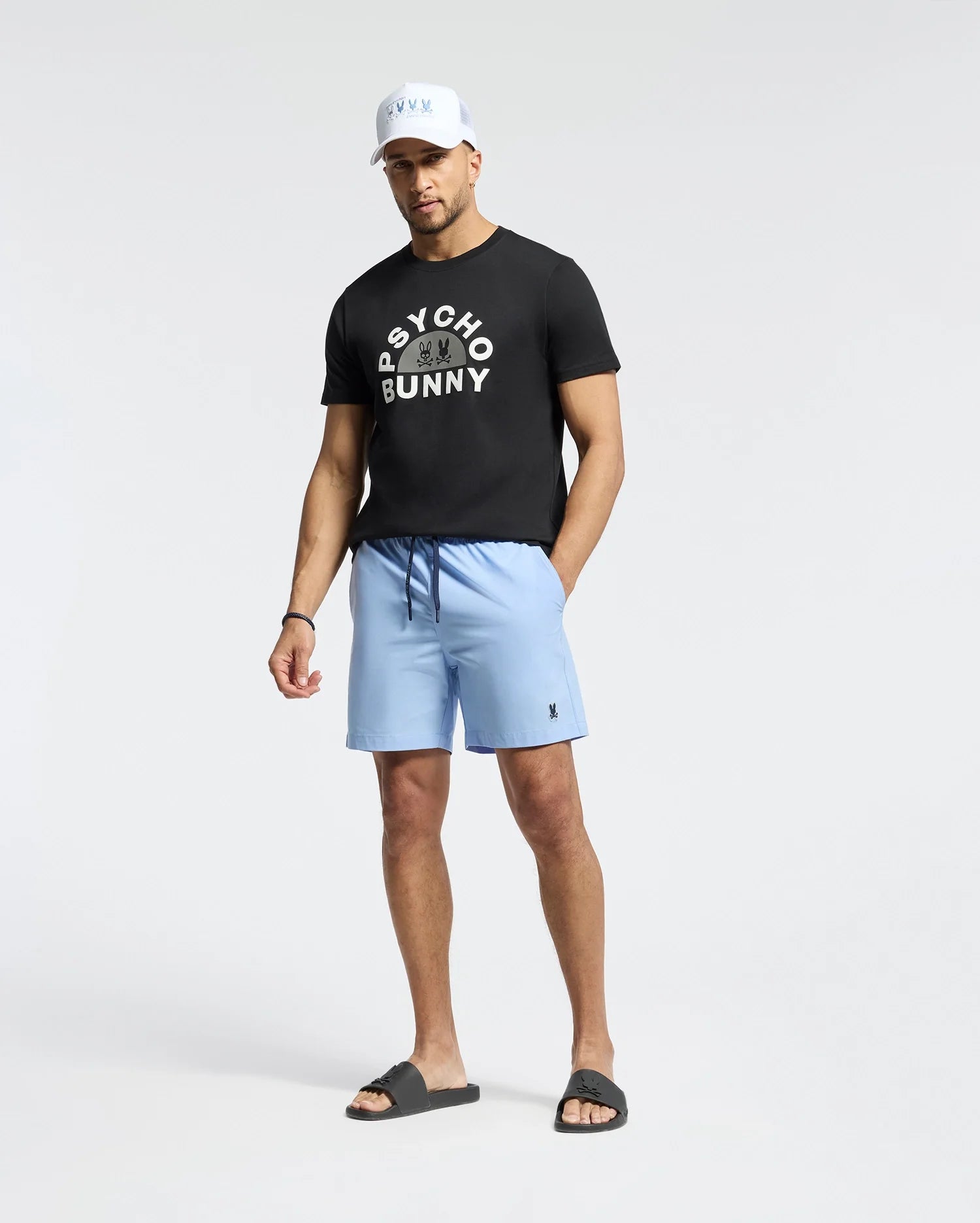 A man stands against a plain backdrop, wearing a black cap and the MENS GOLDIE GRAPHIC TEE by Psycho Bunny, crafted from premium Pima cotton. He's paired it with light blue shorts and black slide sandals, hands by his sides and looking slightly downward.