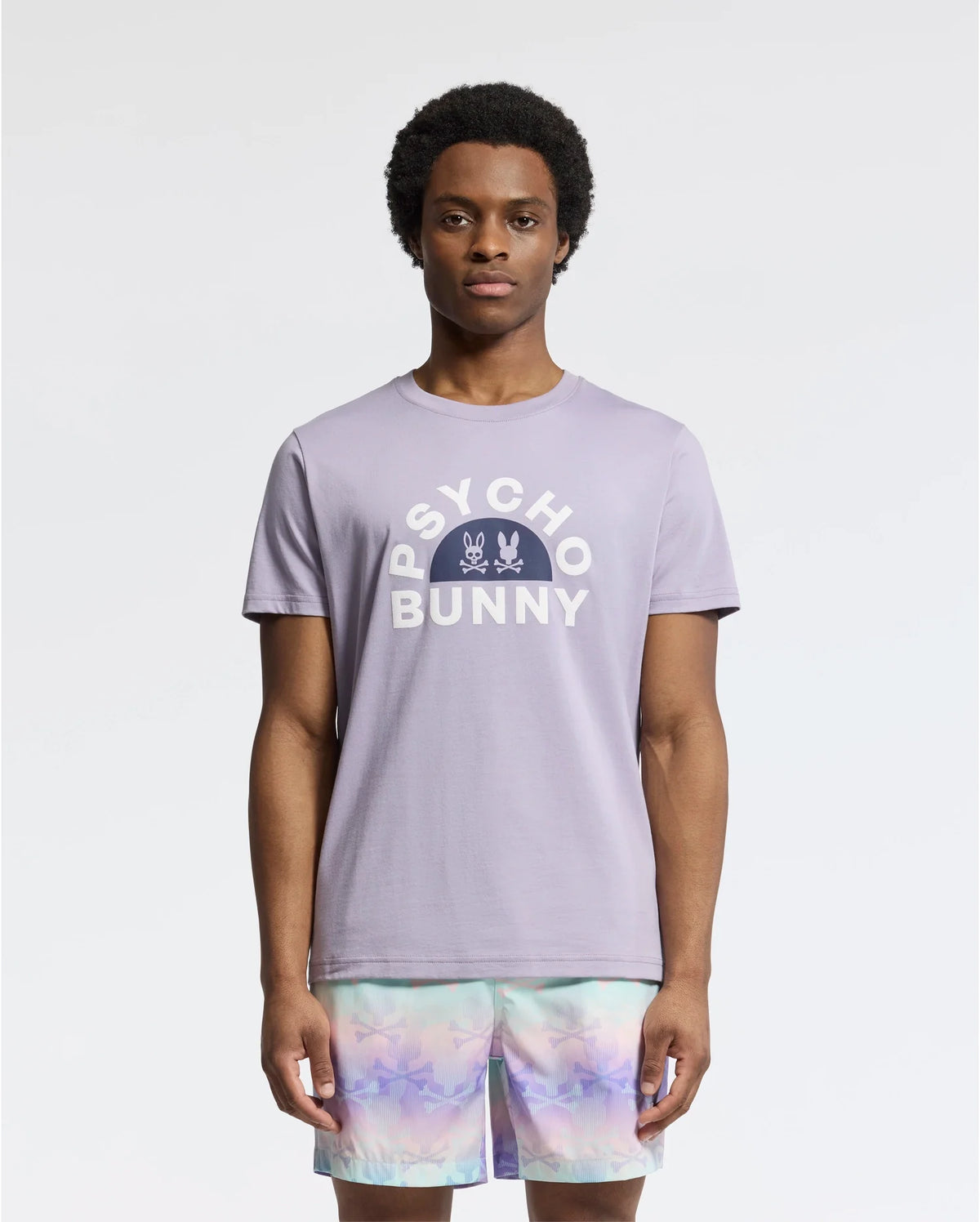 A model displays the MENS GOLDIE GRAPHIC TEE - B6U606F200 by Psycho Bunny, featuring vibrant bunny graphics in luxurious Peruvian Pima cotton, paired with pastel tie-dye shorts on a plain white background.