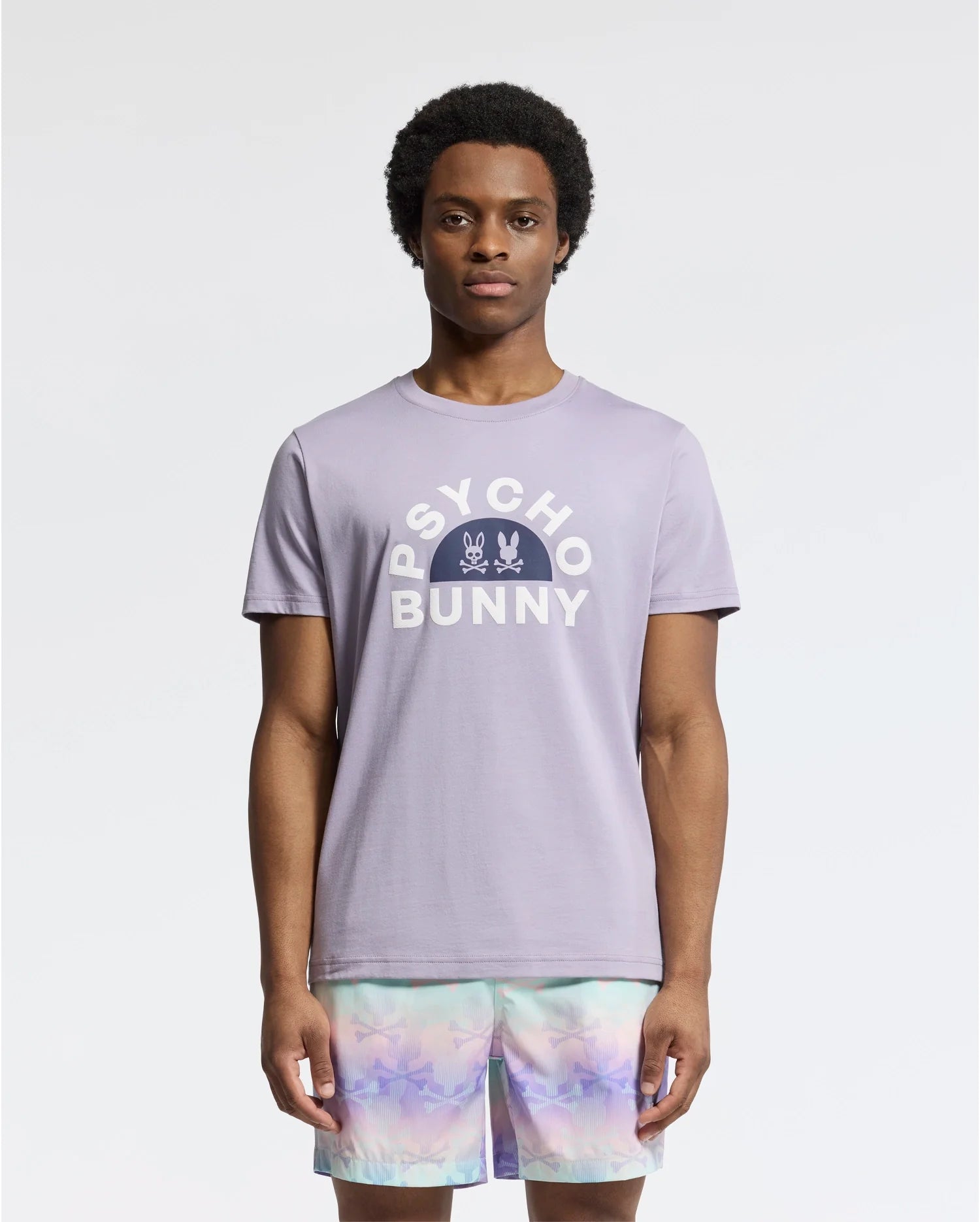 A model displays the MENS GOLDIE GRAPHIC TEE - B6U606F200 by Psycho Bunny, featuring vibrant bunny graphics in luxurious Peruvian Pima cotton, paired with pastel tie-dye shorts on a plain white background.