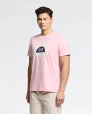 A person is wearing the pink MENS GOLDIE GRAPHIC TEE - B6U606F200 by Psycho Bunny, featuring "PSYCHO BUNNY" in white and a bunny graphic on soft Pima Cotton, paired with light-colored pants against a plain background.