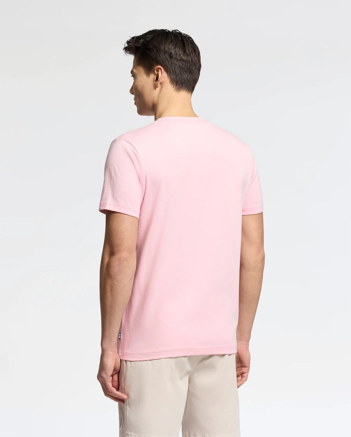 A man, back to the camera, wears a light pink Psycho Bunny MENS GOLDIE GRAPHIC TEE - B6U606F200 made of soft Pima Cotton and beige shorts against a plain white background.
