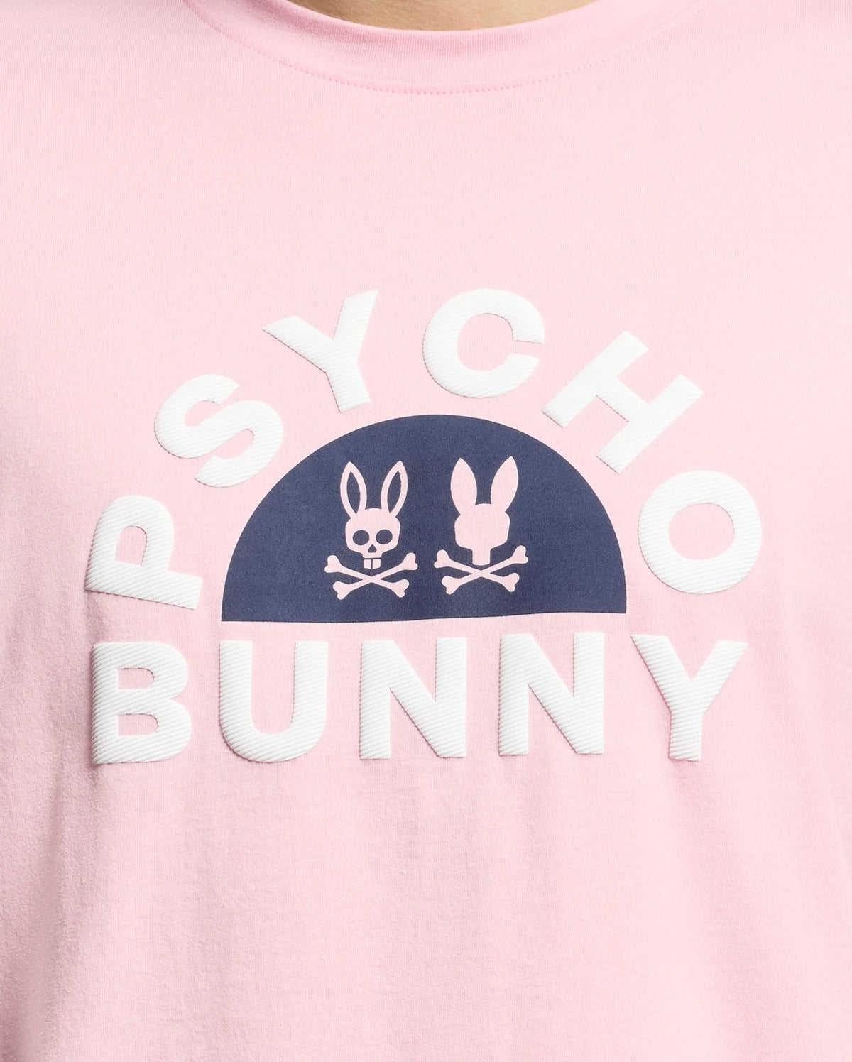 A close-up of the pink Men's Goldie Graphic Tee by Psycho Bunny features white "Psycho Bunny" text around a dark blue half-circle, inside which are two white bunny graphics above crossbones. Crafted from Pima Cotton, it adds a playful edge.