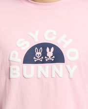 A close-up of the pink Men's Goldie Graphic Tee by Psycho Bunny features white "Psycho Bunny" text around a dark blue half-circle, inside which are two white bunny graphics above crossbones. Crafted from Pima Cotton, it adds a playful edge.