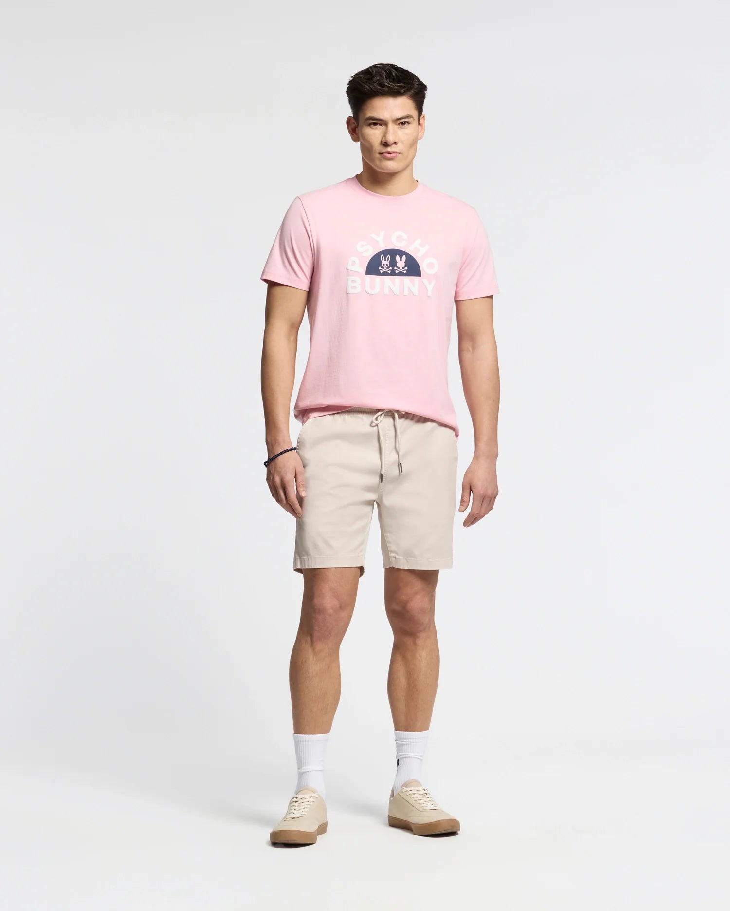A person in a pink "Psycho Bunny" MENS GOLDIE GRAPHIC TEE stands against a white backdrop, pairing it with beige shorts, white socks, and beige sneakers. This ensemble highlights a casual style complemented by the premium comfort of Pima cotton.