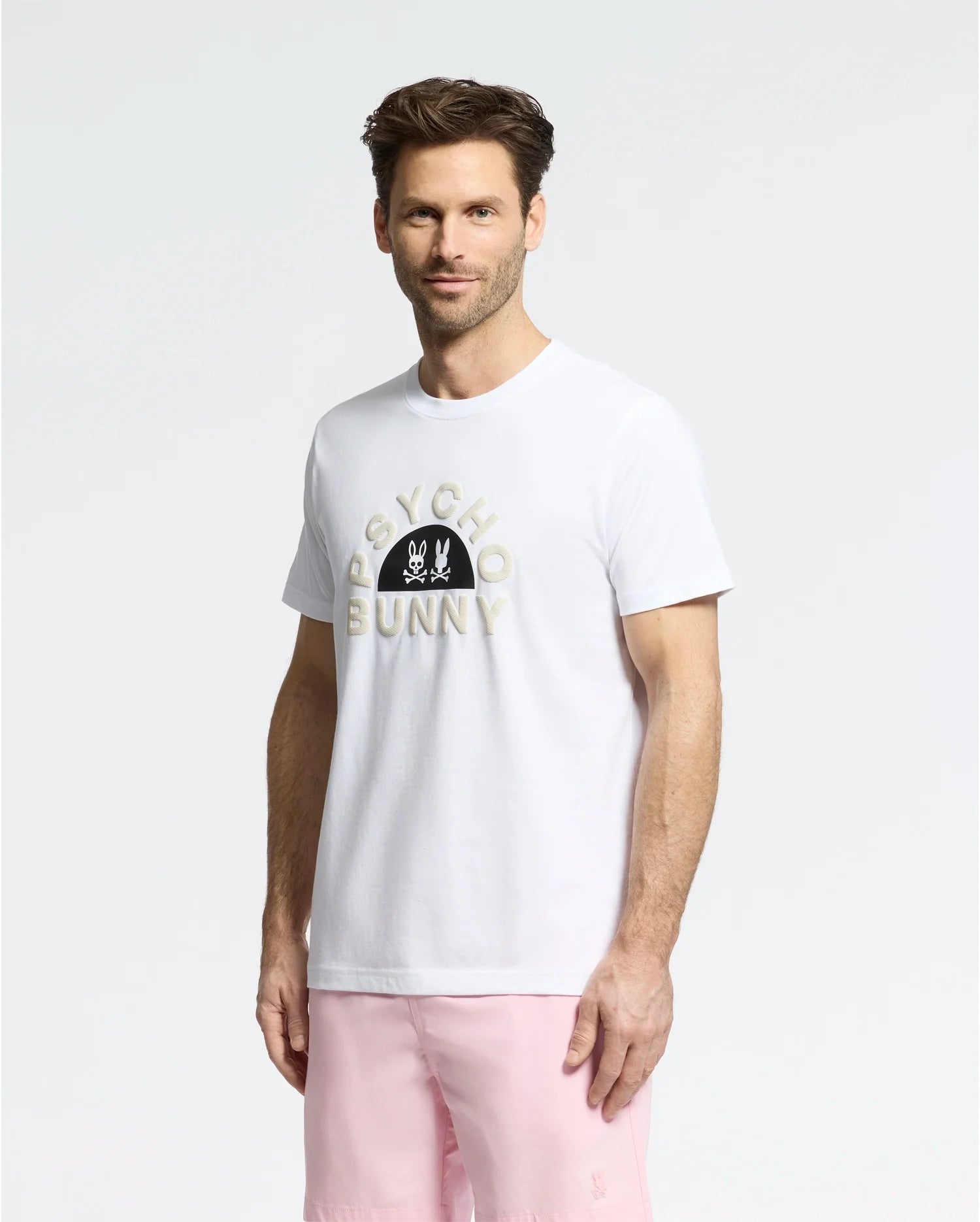 A man confidently poses against a plain background wearing the MENS GOLDIE GRAPHIC TEE (B6U606F200) by Psycho Bunny, featuring the brand’s iconic graphic and logo. His outfit includes light pink shorts, short hair, and a neatly trimmed beard. The T-shirt's Pima cotton ensures comfort and style.