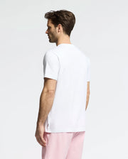 A brown-haired man wears a white Psycho Bunny MENS GOLDIE GRAPHIC TEE - B6U606F200 with a subtle bunny graphic, paired with pink pants. He stands with his back turned, looking left against a plain, light background.