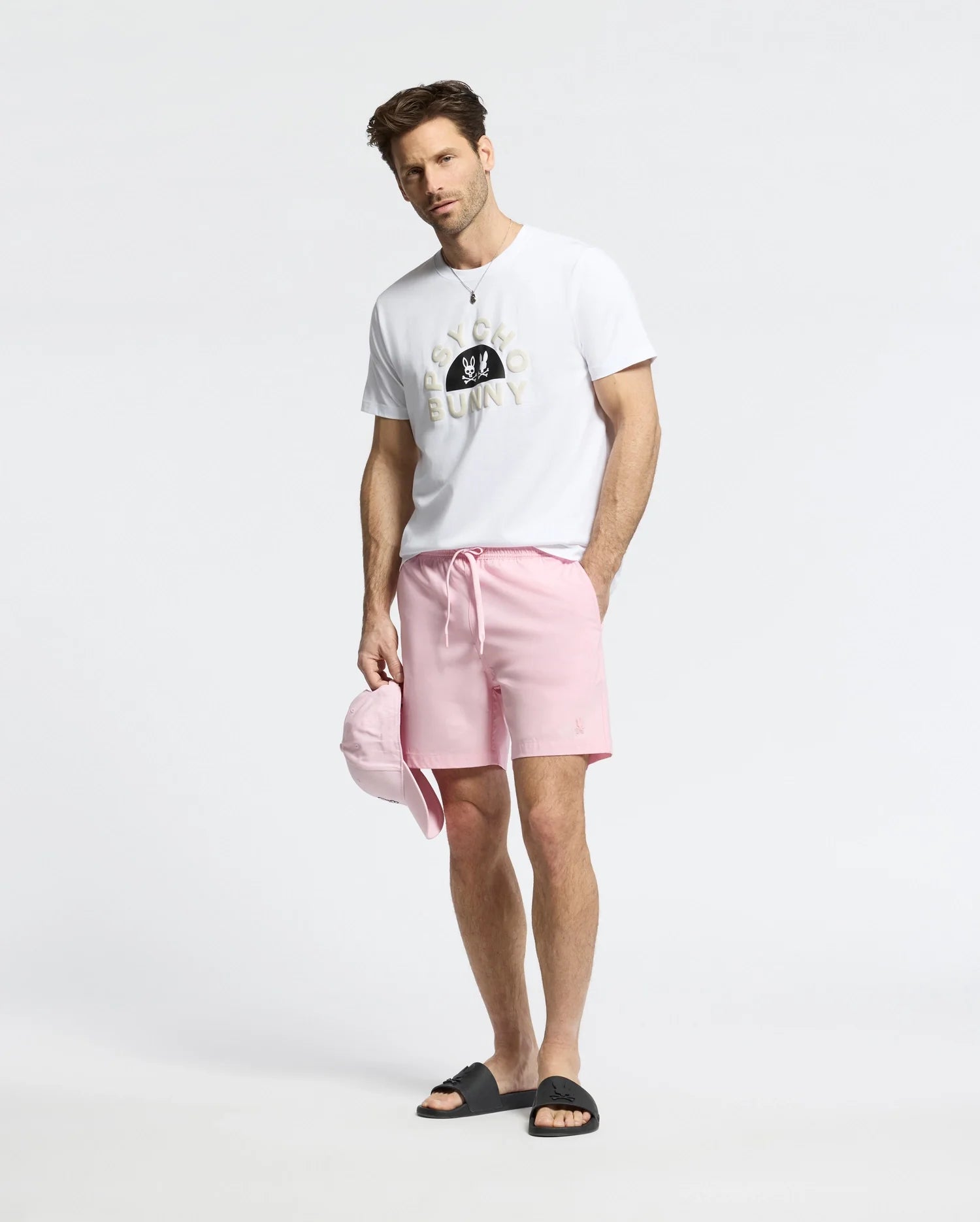 A man in a bright setting wears Psycho Bunny’s white MENS GOLDIE GRAPHIC TEE, light pink shorts, and black slide sandals. He holds a matching pink cap while looking at the camera.