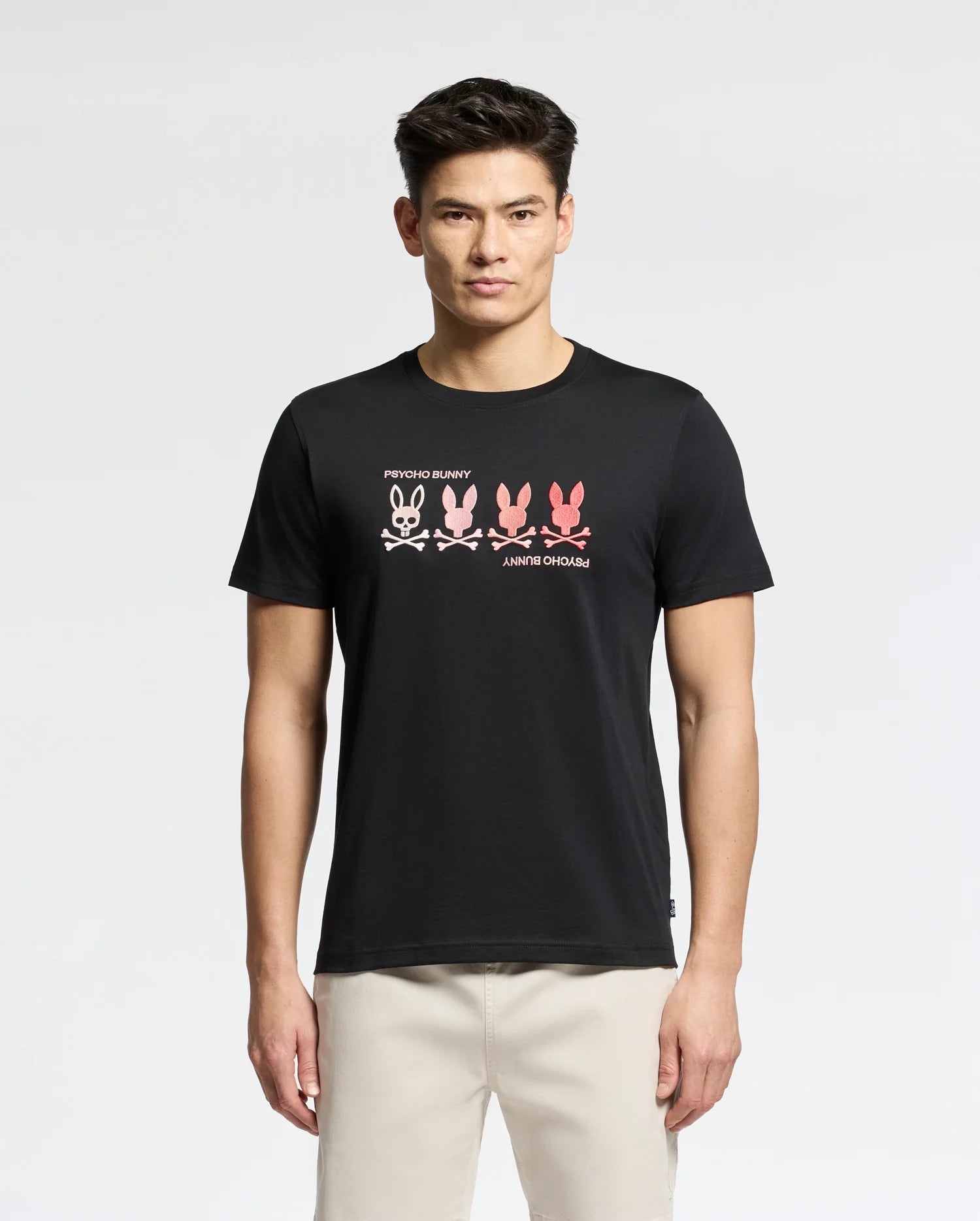 Someone stands against a plain background wearing a Psycho Bunny MENS OCEANIA EMBROIDERED GRAPHIC TEE. The black Pima Cotton shirt features an embroidered bunny graphic with four gradient bunnies (white to pink), "RHYTHM BUNNY" above, and "ANRHY OHOMAJ" below, exuding retro vibes.