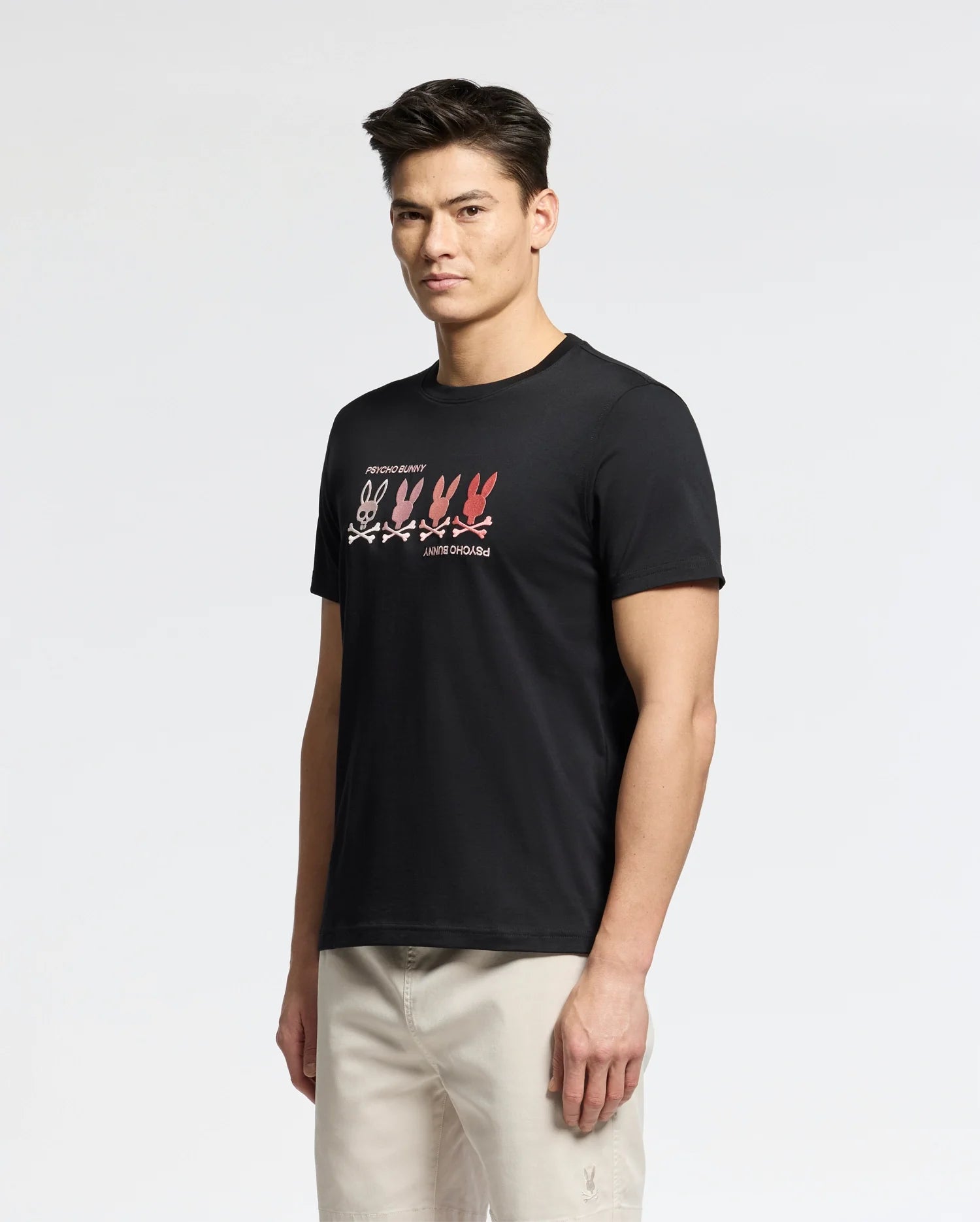A person wearing Psycho Bunny's MENS OCEANIA EMBROIDERED GRAPHIC TEE - B6U607F200 stands against a plain white background. The black Pima cotton tee features retro embroidery with pink and red bunny outlines, while the person wears beige pants and looks toward the camera.