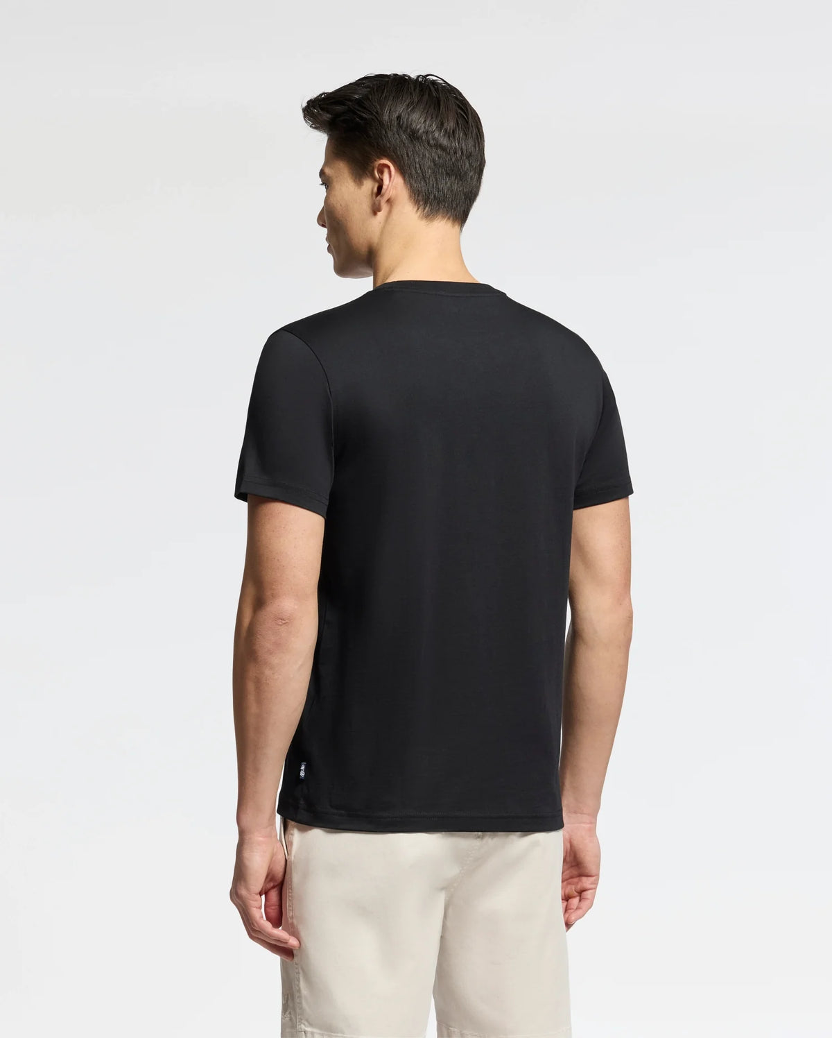 A person faces away from the camera wearing a black Psycho Bunny MENS OCEANIA EMBROIDERED GRAPHIC TEE - B6U607F200, paired with light-colored shorts. The plain, neutral background lends a subtle retro vibe.