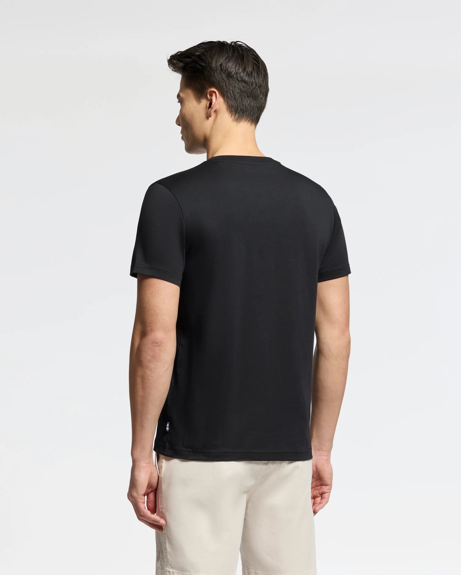 A person faces away from the camera wearing a black Psycho Bunny MENS OCEANIA EMBROIDERED GRAPHIC TEE - B6U607F200, paired with light-colored shorts. The plain, neutral background lends a subtle retro vibe.