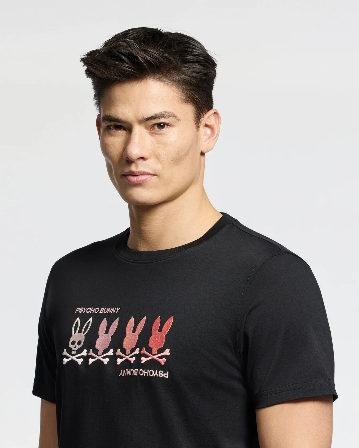 A person with short dark hair is wearing a black Psycho Bunny MENS OCEANIA EMBROIDERED GRAPHIC TEE, showcasing four bunny skulls in gradient colors from pink to red for a retro feel. "Psycho Bunny" is embroidered around the design on a light-colored background.