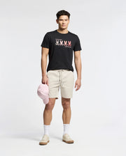 A man stands against a plain background wearing a Psycho Bunny black Pima Cotton Tee featuring an embroidered bunny, beige shorts, white socks, and light brown sneakers. He holds a light pink baseball cap at his side.