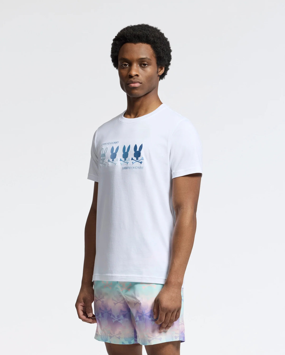 The person poses against a plain background, wearing a Psycho Bunny MENS OCEANIA EMBROIDERED GRAPHIC TEE - B6U607F200 with bunny embroidery and multicolored shorts. Their short hair enhances the retro vibe as they gaze directly at the camera.