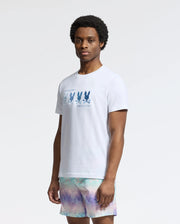 The person poses against a plain background, wearing a Psycho Bunny MENS OCEANIA EMBROIDERED GRAPHIC TEE - B6U607F200 with bunny embroidery and multicolored shorts. Their short hair enhances the retro vibe as they gaze directly at the camera.