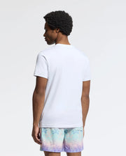 A person with curly hair stands facing away, wearing a Psycho Bunny Men's Oceania Embroidered Graphic Tee and pastel shorts. The plain, light-colored background enhances the retro vibes of the effortlessly cool scene.