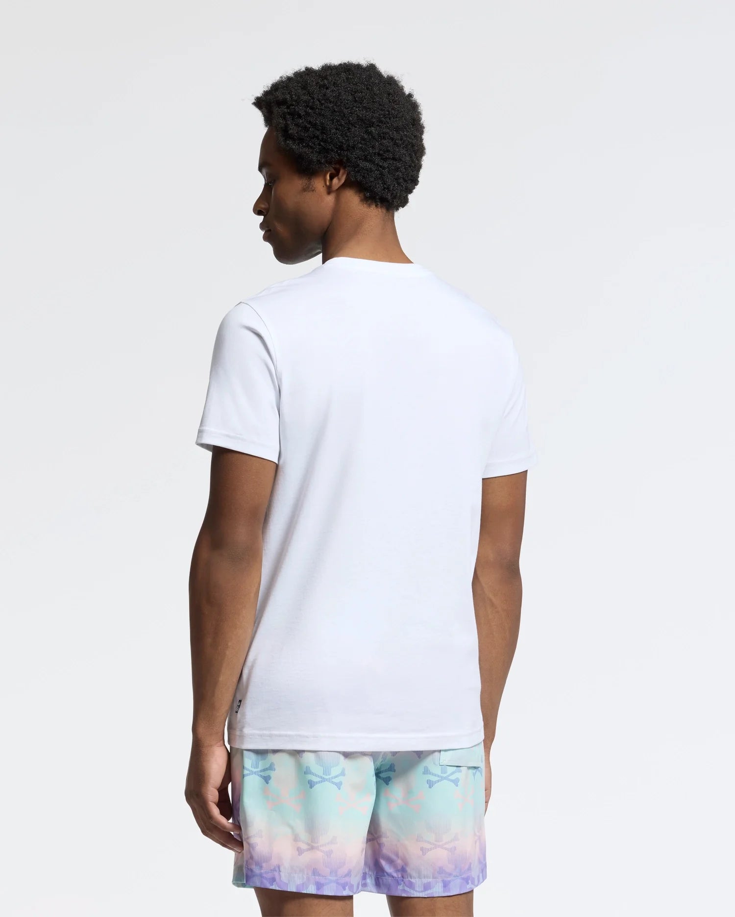 A person with curly hair stands facing away, wearing a Psycho Bunny Men's Oceania Embroidered Graphic Tee and pastel shorts. The plain, light-colored background enhances the retro vibes of the effortlessly cool scene.