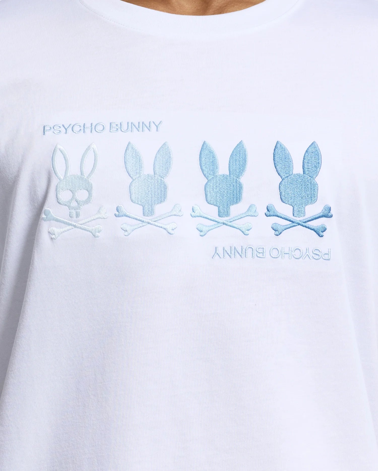 The MENS OCEANIA EMBROIDERED GRAPHIC TEE by Psycho Bunny is a white Pima cotton shirt featuring blue "Psycho Bunny" text with embroidered bunny silhouettes over crossed bones, offering retro vibes with a light-to-dark blue gradient effect.