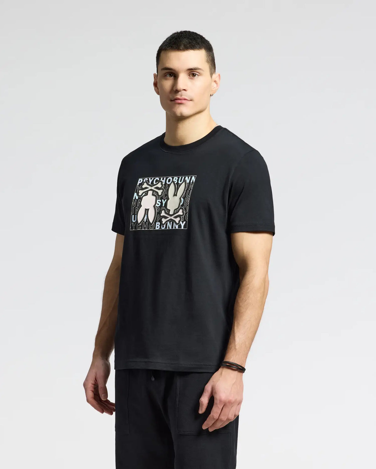 A man stands against a plain background in a Psycho Bunny's MENS GOLD COAST GRAPHIC TEE - B6U631F200, made from Pima cotton. The black T-shirt features rabbits and text. He has short dark hair, wears dark pants and a wristband on his left wrist, embodying classic fit with premium craftsmanship.