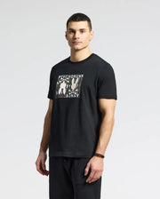 A man stands against a plain background in a Psycho Bunny's MENS GOLD COAST GRAPHIC TEE - B6U631F200, made from Pima cotton. The black T-shirt features rabbits and text. He has short dark hair, wears dark pants and a wristband on his left wrist, embodying classic fit with premium craftsmanship.