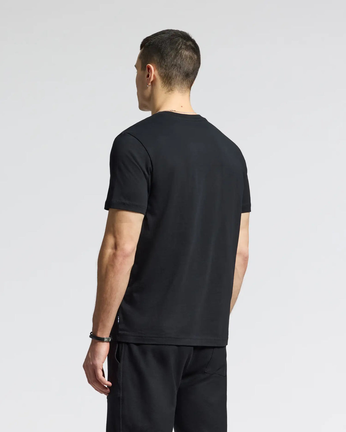 A person with short hair, back to camera, wears a Psycho Bunny MENS GOLD COAST GRAPHIC TEE - B6U631F200 and shorts, their classic fit highlighted by a plain, light backdrop that hints at premium craftsmanship.