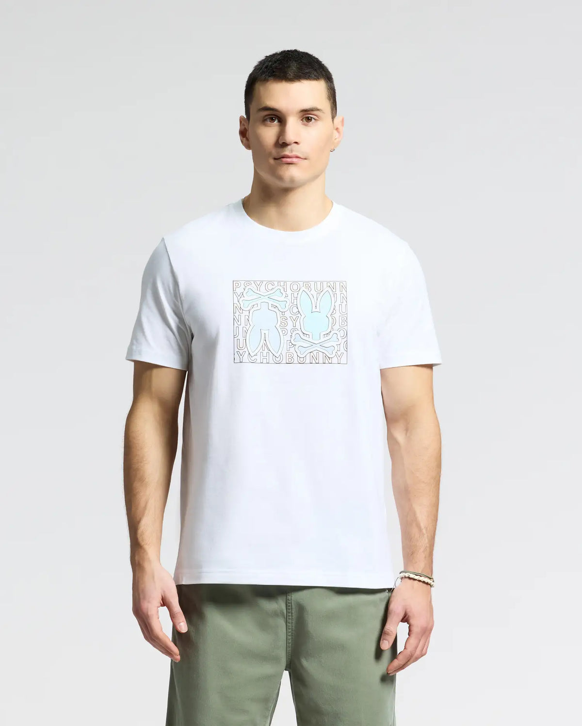 A person is wearing a Psycho Bunny MENS GOLD COAST GRAPHIC TEE - B6U631F200, a premium Pima cotton white t-shirt with abstract figures in a square, paired with green pants. The classic fit enhances the simple light background elegantly.