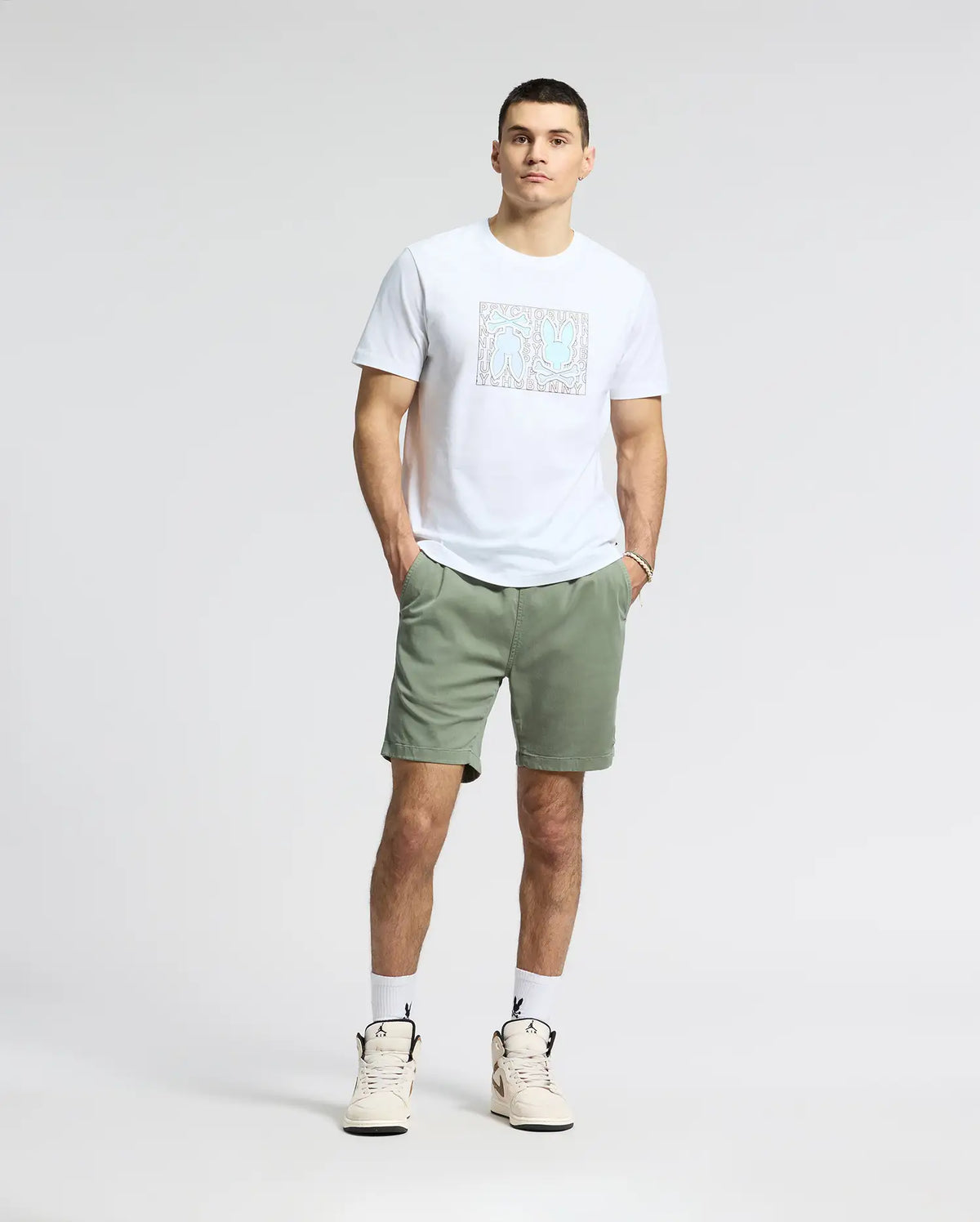 A man stands against a plain background wearing a white Psycho Bunny MENS GOLD COAST GRAPHIC TEE - B6U631F200, green shorts, and white sneakers, with his hands in his pockets and short hair.